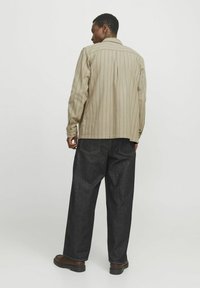 Camicetta a righe beige a maniche lunghe con polsini a bottoni, indossata con pantaloni baggy in denim scuro e scarpe marroni. Sfondo neutro, focalizzato sui dettagli dell'abbigliamento.