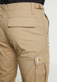 Beiget väriset, kestävästä kankaasta tehdyt työhaarukkaiset housut, joissa on useita taskuja, sivulla oleva logopatch ja näkyvä ompelus.