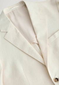 Gros plan sur un blazer crème montrant un revers cranté, une poche poitrine, une doublure intérieure et un unique bouton marron à l'avant.