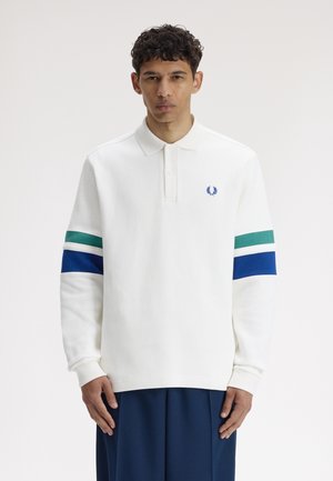 Junger Mann trägt ein weißes, langärmliges Polohemd mit grünen und blauen Streifen an den Ärmeln, steht vor einem schlichten hellen Hintergrund.