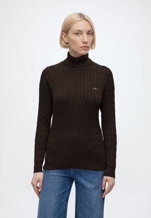 Blonde Frau mit goldenen Creolen, einem dunkelbraunen Zopfmuster-Rollkragenpullover und blauen Jeans, vor einem einfarbigen weißen Hintergrund stehend.