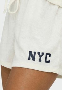Weiße Frottee-Shorts mit elastischem Bund und Kordelzug, versehen mit auffälligem navyblauen "NYC"-Schriftzug am unteren Saum. Weicher Stoff und lässige Passform.