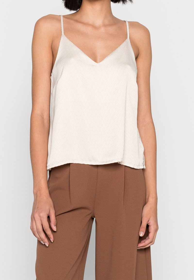 Femme portant un haut sans manches texturé blanc à col en V avec fines bretelles et un pantalon marron ample sur un fond uni.