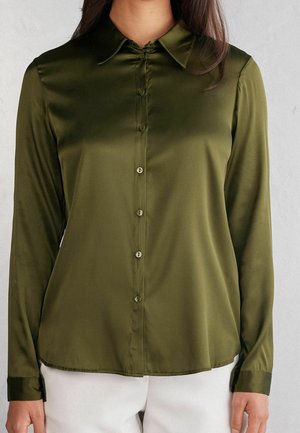 Kobieta ubrana w oliwkowozieloną satynową bluzkę z długim rękawem, z guzikami i kołnierzykiem, w połączeniu z białymi spodniami, widziana od szyi do bioder.