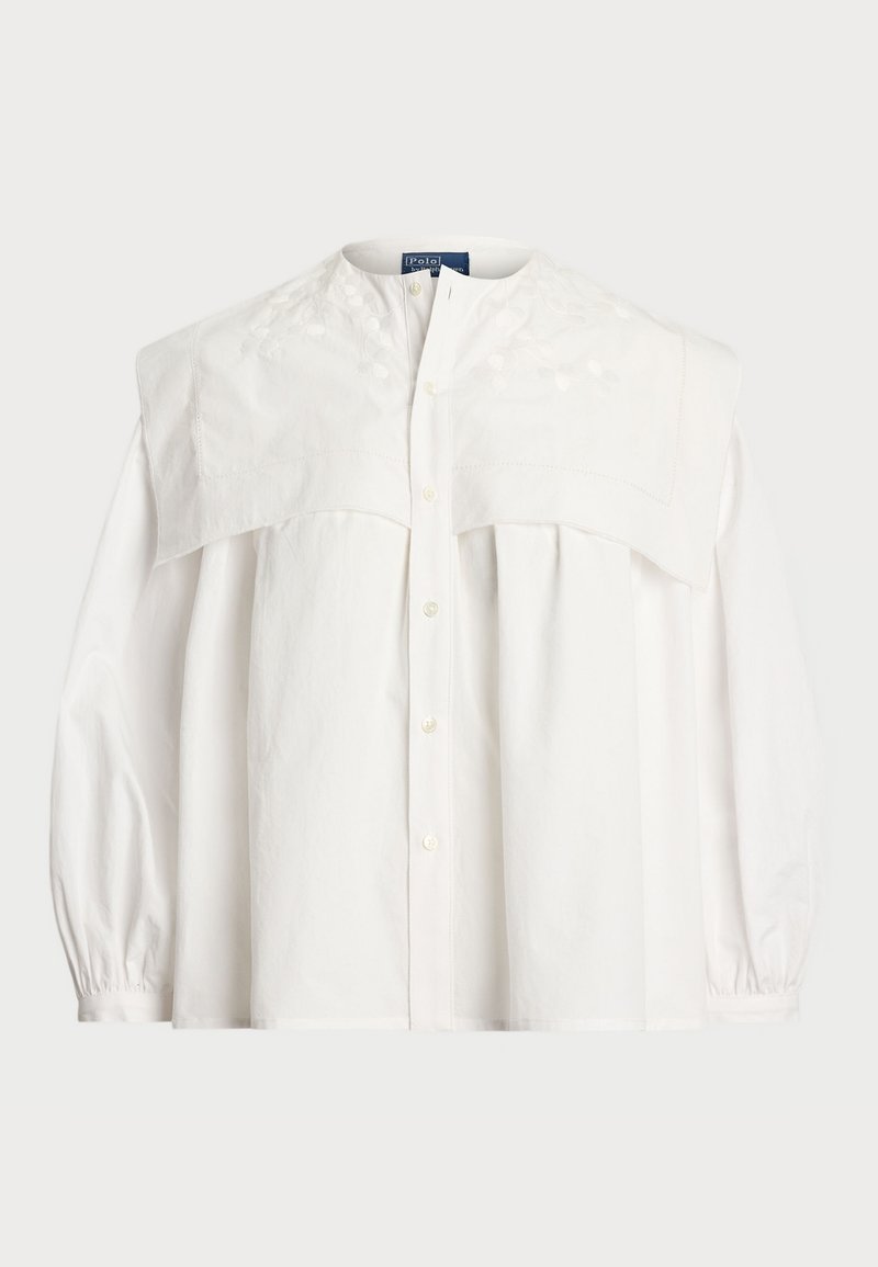 Blouse en coton blanc avec un large empiècement brodé, fermeture à boutons sur le devant, et manches bouffantes avec des poignets élastiques.