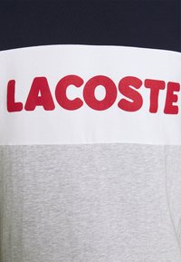 Bavlnené tričko s vodorovnými farebnými blokmi: námornícka modrá, biela a svetlo šedá. Veľký červený text "LACOSTE" je výrazne zobrazený.