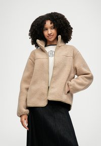 Beige fleece zip-up jacket with a high collar, soft texture, and contrasting zippers; worn over a light-colored top and dark skirt.