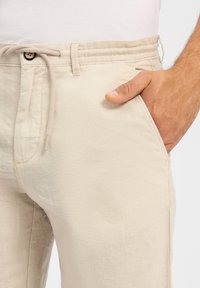 Beige Leinenhosen mit einem verstellbaren Bund, Knopfverschluss und Seitentasche, glatter Textur und lockerer Passform.