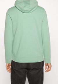 Persona vestida con una sudadera verde clara y pantalones de pana negros, vista desde atrás con las manos relajadas a los lados.