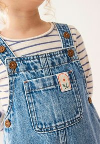 Pantalones de mezclilla con botones de madera, tejido en azul claro con una textura sutil. Incluye un parche con un diseño floral en el bolsillo y una parte superior a rayas.
