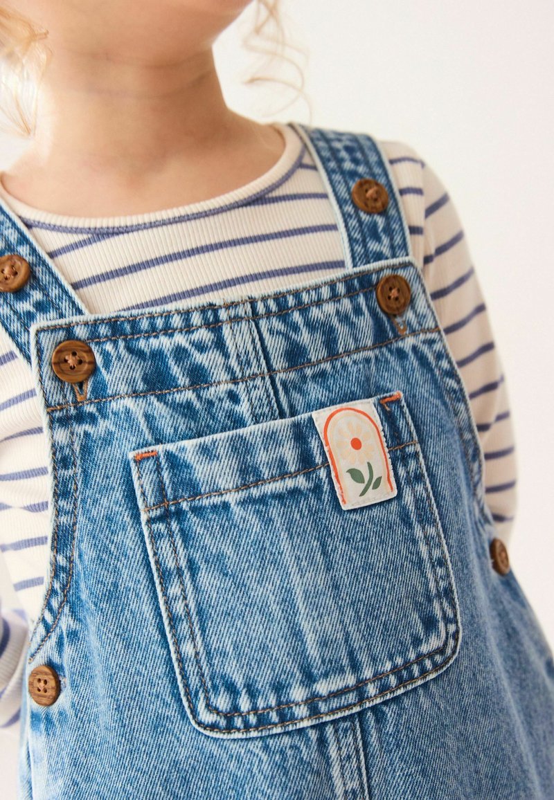 Pantalones de mezclilla con botones de madera, tejido en azul claro con una textura sutil. Incluye un parche con un diseño floral en el bolsillo y una parte superior a rayas.