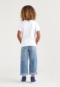 Bambino con capelli ricci che sta in piedi di spalle indossando una maglietta bianca, jeans blu arrotolati, calze a righe e sneakers alte blu.