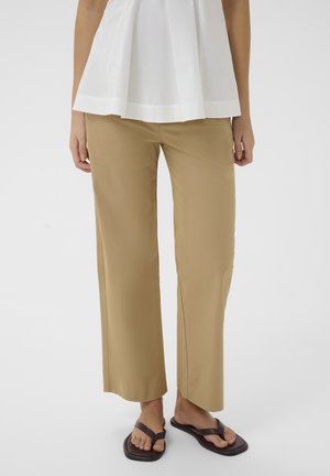 Osoba ubrana w beżowe spodnie o szerokich nogawkach, biały bezrękawnik i ciemnobrązowe sandały z kwadratowym noskiem, stoi na jednolitym tle.