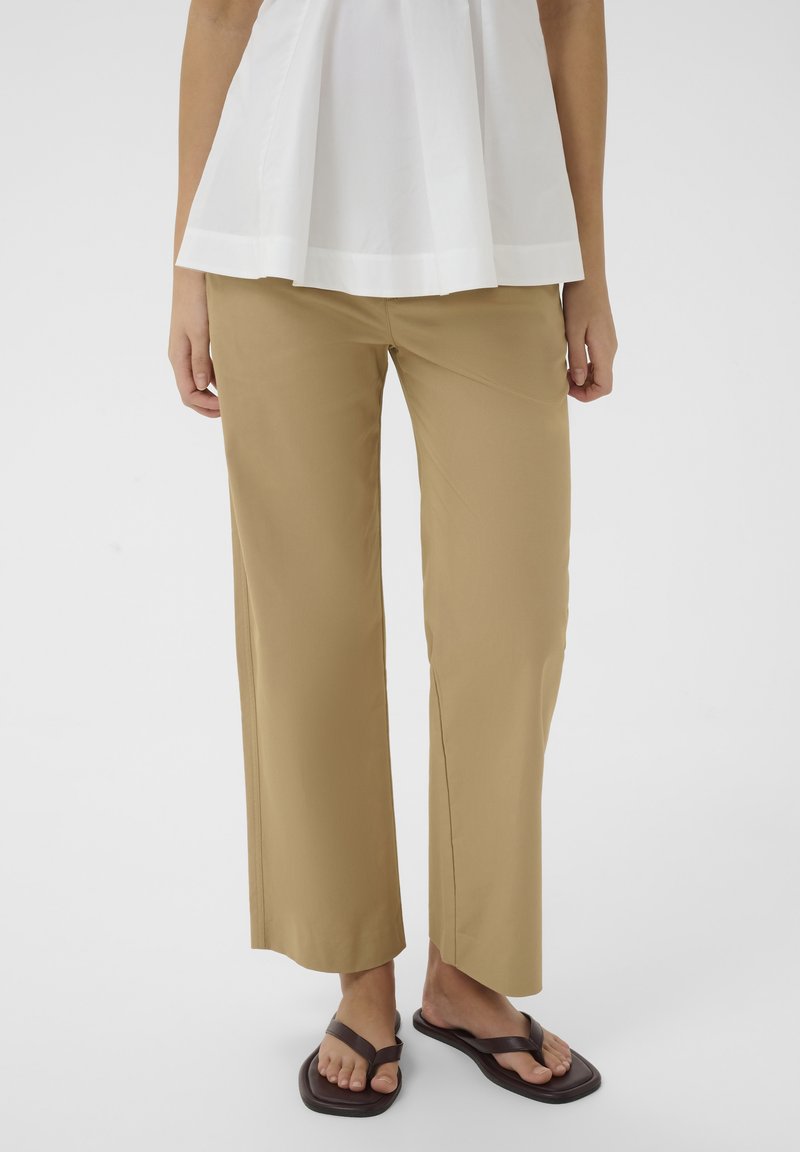 Personne portant un pantalon beige à jambes larges, un haut blanc sans manches et des sandales marron foncé à bout carré, debout devant un fond uni.