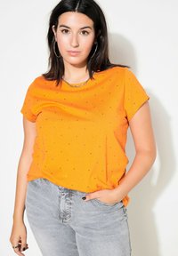 Orange t-shirt med kort ärm, små svarta dekorationer, rund halsringning och en avslappnad passform, i kombination med ljusgrå jeans.