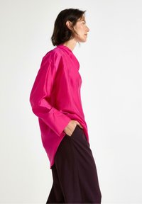 Fuchsia langærmet bluse i et glat stof, med høj halsudskæring og løs pasform, parret med mørke vidde bukser.