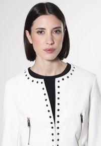 Giacca in pelle bianca con dettagli in rivetti neri lungo il colletto, una tasca con zip frontale e una texture liscia e lucida.