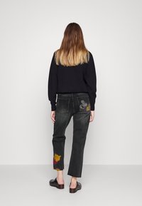 Pull en maille noire, jean gris foncé avec des personnages brodés sur les poches arrière, ourlet court, et sabots à talon bloc et dos ouvert.