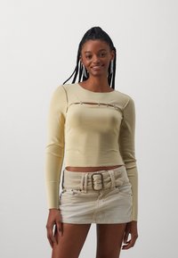 Ljusgul långärmad topp med en figurnära design, som har en horisontell skärning med tre dekorativa knappar. Bärs med en beige kordflors mini kjol.