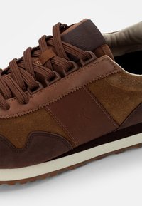 Brun sneakers med en blandning av slätt läder och mocka. Har runda skosnören, vadderad krage och en vit gummiyttersula med texturerade detaljer.