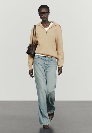 Kobieta w oversize beżowym swetrze na zamek, jasnoniebieskich dżinsach z szerokimi nogawkami, ciemnych okularach przeciwsłonecznych, niosąca brązową torebkę na ramię, spacerująca po szarej podłodze.