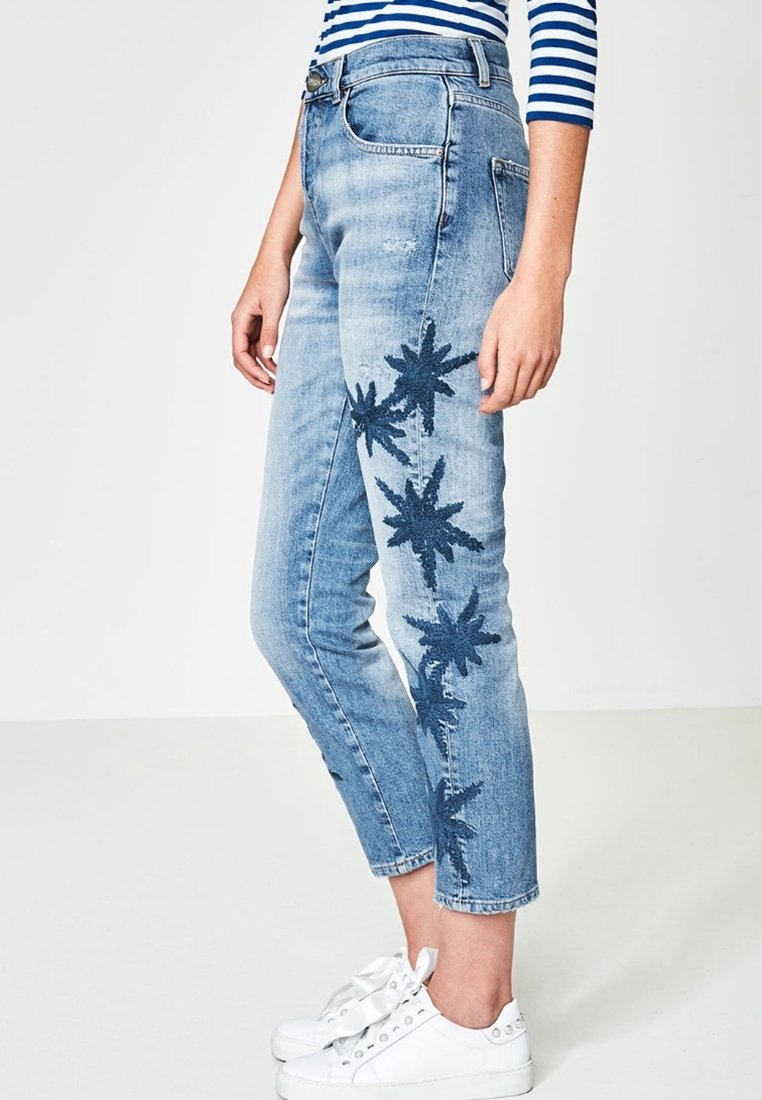 Jean court bleu clair avec broderie florale en forme d'étoile bleu foncé le long de la jambe extérieure, porté avec des baskets blanches et un haut rayé.