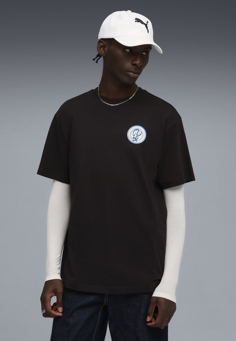 Sort bomulds T-shirt med korte ærmer, prydet med et blåt og hvidt cirkulært logo. Kombineret med en hvid kasket med et sort logo.