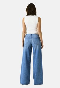 Vrouw met schouderlang donker haar, gekleed in een witte mouwloze top en een wijde blauwe jeans, gezien van achteren tegen een effen achtergrond.