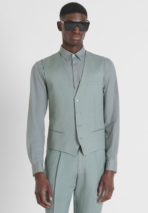 Gilet vert clair avec une chemise assortie, présentant un design épuré, une texture lisse et cinq boutons. Le modèle porte des lunettes de soleil sombres et angulaires.