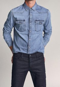 Camicia di denim azzurro chiaro con chiusura con bottoni, due tasche sul petto e rifinitura a contrasto. Abbinata a jeans in denim scuro.