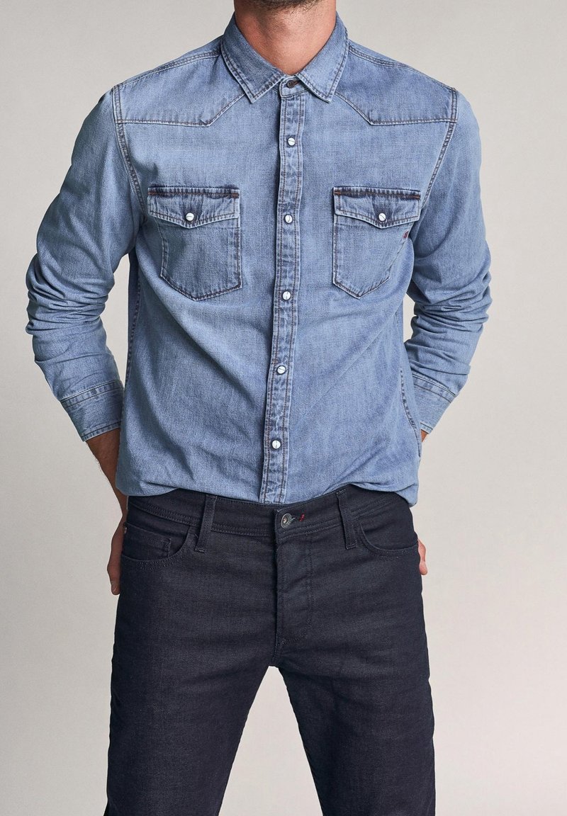 Camicia di denim azzurro chiaro con chiusura con bottoni, due tasche sul petto e rifinitura a contrasto. Abbinata a jeans in denim scuro.