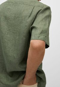 Zielona lniana koszula z krótkim rękawem o luźnym kroju, z tylnym karczkiem i widoczną fakturą. Nosi się ją na jasnych spodniach.