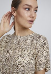 Top s leopardím vzorem, který má kulatý výstřih a krátké rukávy, vyrobený z lehkého materiálu v béžové a hnědé barevné paletě.