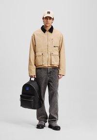 Beige lerretjakke med svart krage, to frontlommer, båret over en tan topp. Mørkegrå denimjeans, svart ryggsekk med blå detaljer.