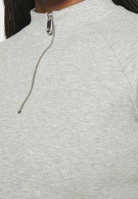 Primo piano di un top in tessuto grigio a costine con una cerniera argento parzialmente chiusa al collo, indossato da una persona con capelli scuri.