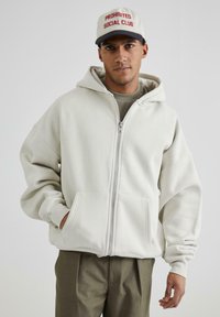 Felicemente indossato su una maglietta grigia, la felpa bianca con zip è realizzata in un materiale morbido e presenta un cappuccio ampio e tasche frontali, abbinata a pantaloni color khaki.