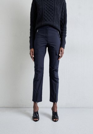 Woman wearing black cable knit sweater, black cargo pants with zipper details, and black open-toe heeled shoes, standing against white wall.