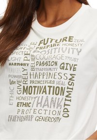 Bílý bavlněný tričko s typografickým designem a motivačními slovy v různých písmových stylech, převážně v zelené a stříbrné barvě.