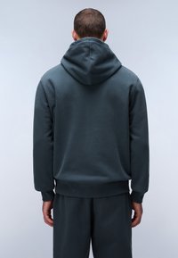 Sudadera en un tono verde azulado oscuro con una gran capucha ajustable, puños y cintura de canalé. Textura de tela suave, diseñada con un corte relajado.
