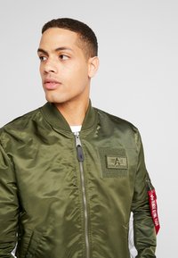 Olivgrüne Bomberjacke aus glänzendem Nylonmaterial mit Reißverschluss vorne, geripptem Kragen und einem Patch auf der Brust. Mit einem roten Etikett am Ärmel.