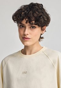 Ljus krämfärgad sweatshirt med rund halsringning, raglanärmar och ett broderat "POLO CLUB" i guld på bröstet. Slät textur.