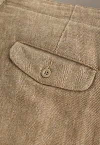 Tissu en chevron beige et blanc avec une poche à rabat boutonnée, présentant un bouton beige à quatre trous et des détails de couture visibles.