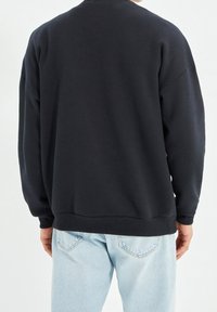 Sudadera negra de corte relajado, con cuello redondo y puños acanalados, combinada con jeans azul claro, mostrando la vista posterior.