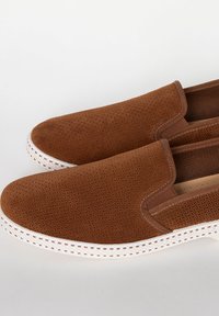 Bruine instap schoenen met geperforeerde textuur, witte rubberen zool en contrasterende stiksels langs de randen.