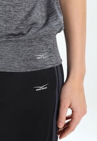 Persona vistiendo pantalones deportivos oscuros y una camiseta gris, ambos con logotipos de "Venice Beach" cerca de la cintura, con la mano relajada a su lado.