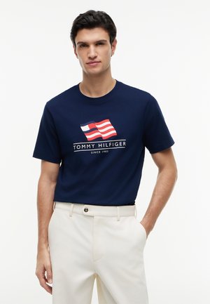 Άνδρας που φοράει μπλε ναυτικό μπλουζάκι Tommy Hilfiger με λογότυπο σημαίας σε κόκκινο, λευκό και μπλε και λευκό παντελόνι, στέκεται με το ένα χέρι στην τσέπη.