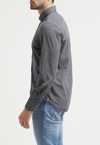 Harmaa napillinen paita, jossa on tekstuuri ja kaulus sekä pitkät hihat, yhdistetty sinisiin farkkuihin.