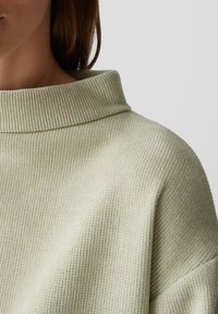 Frau, die einen hellgrünen gerippten Strickpullover mit hohem, gefaltetem Kragen trägt, von Hals bis Schulterbereich vor einem schlichten Hintergrund gezeigt.