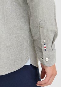 Grå skjorte med lange ermer, med hvite knapper, skrå mansjetter og en triangular detalj med rød, hvit og blå striper.