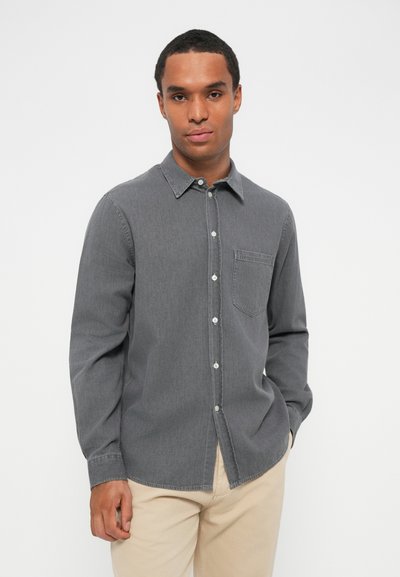 Chemise grise à manches longues en tissu doux, avec un col boutonné, une poche avant et des boutons blancs. Associée à un pantalon de couleur neutre.